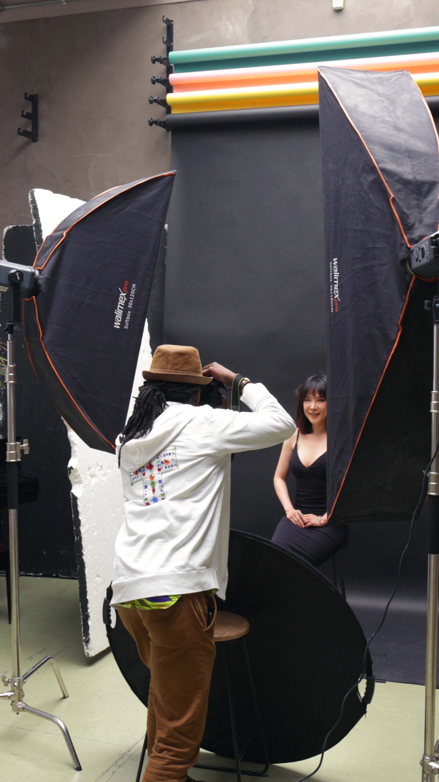 In a photography studio, a Black photographer is capturing an image of a smiling East Asian woman. The woman sits confidently, her expression radiating joy and warmth.