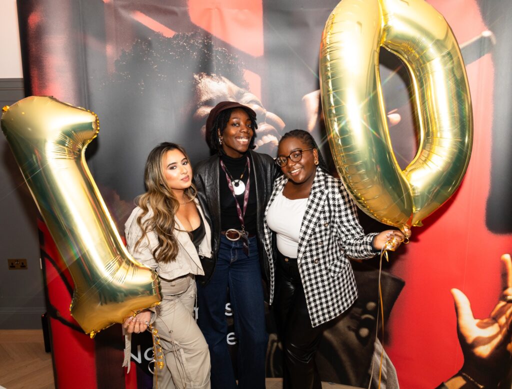 Mati, a young Black woman wearing a brown cap, black turtleneck, black leather jacket, red belt, and jeans, stands at the center of the photo with two female friends beside her. They are holding large silver "1" and "0" foil balloons, celebrating the Be United "10 Years of History" event. The group stands beside a Be United banner, smiling together in front of a vibrant backdrop.