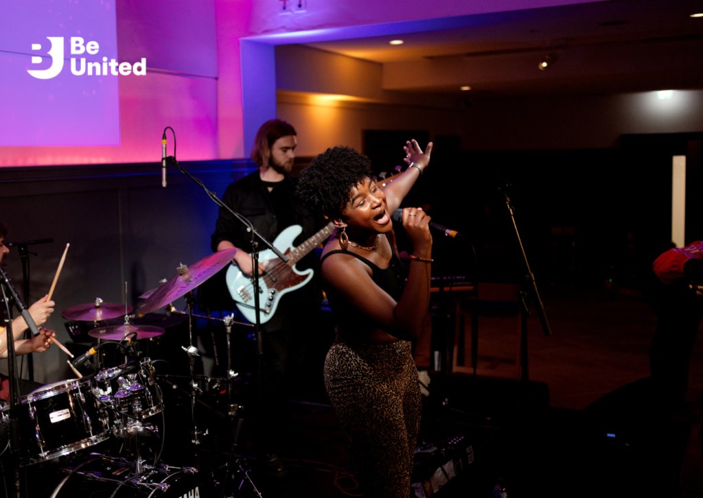 A Black woman with short hair, wearing a black top and leopard print trousers, passionately singing on stage while holding a microphone. A drummer and bassist are visible in the background.