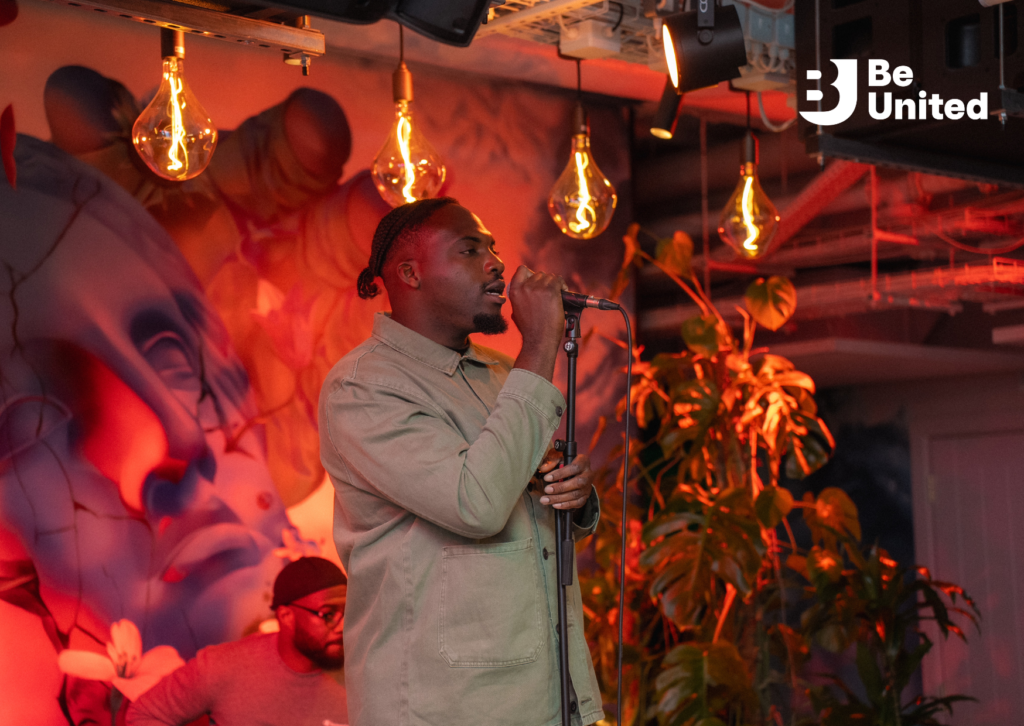 Ay Bagz, a Black man in a beige shirt-jacket, standing with a microphone in a mic stand, singing. Light fixtures, a modern mural of an ancient statue, a plant, and a guitarist are visible in the background.