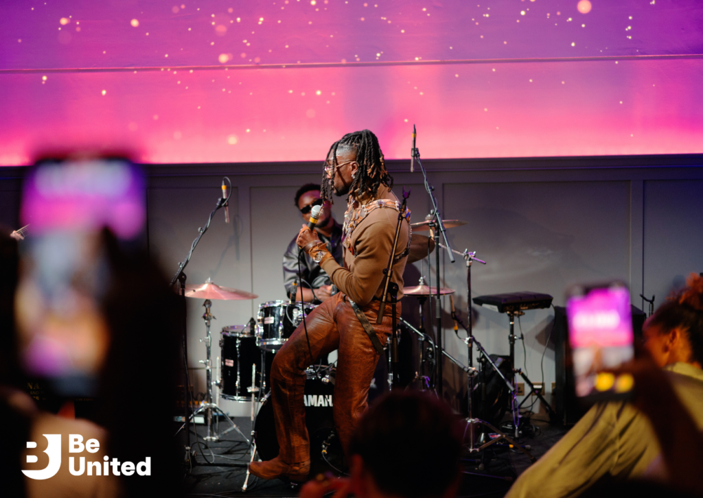 A Black man with long locks, wearing a brown top, brown leather trousers, and cowboy boots, singing on stage. A Black drummer is visible in the background. People in the foreground are holding up their smartphones.