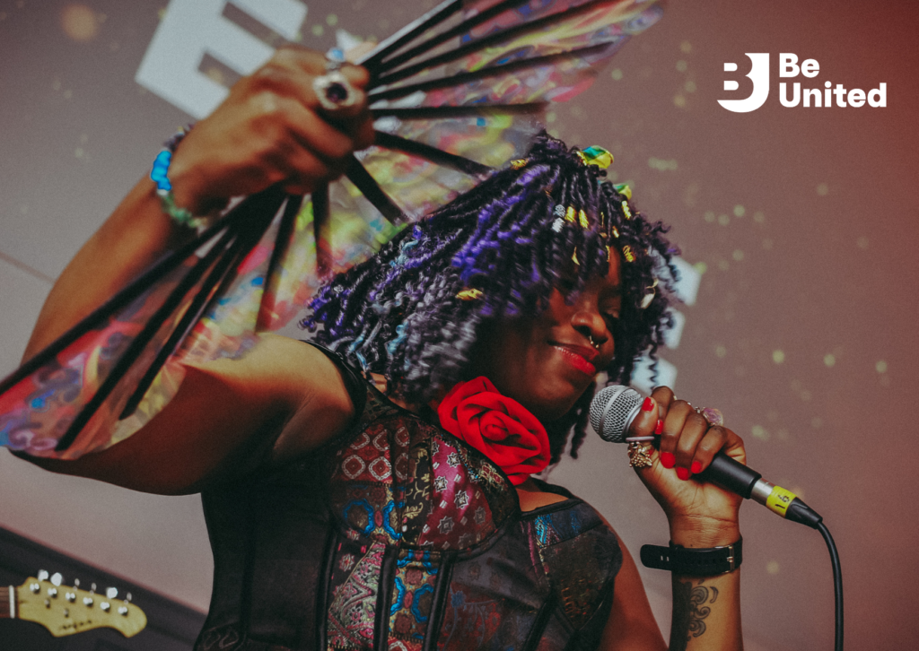 Eyve, a Black woman with black and purple curly hair, wearing a multi-colored dress and a red rose necklace, holding a microphone and smiling. She accessorizes with a hand fan and red lipstick.