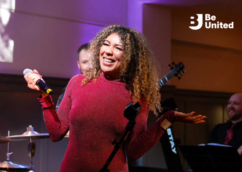 A fair-skinned Black woman (Ziggy) with curly light-brown hair, wearing a shiny pink jumpsuit, standing on stage and holding a microphone, smiling.