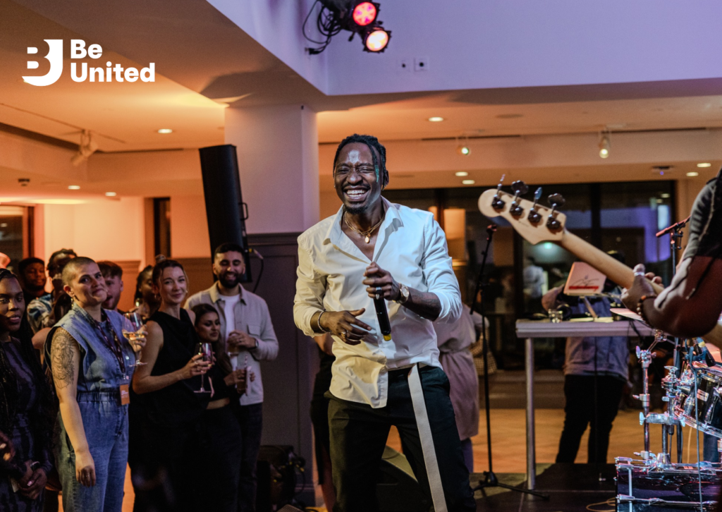 A Black man in a White T-Shirt and Black jeans (Sean Focus) on stage, smiling and holding the mic. The crowd and the band is seen in the foreground/background. 