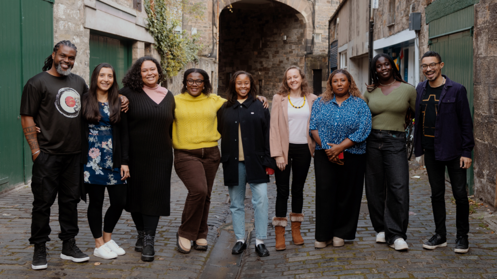 A group photo of some of Be United's dedicated Operational team and Board members (9 people) standing outside in a line, from left to right: Collin Hills (Black man in a Black T-shirt and jeans), Zarine Manekshaw (South Asian woman with dark long straight hair in a flowery dress), Sharon Thomas (Black woman with wavy hair, in a Black dress and boots), Dr. Thelma Okey-Adibe (Black woman in a yellow sweater and brown trousers), Kassana Garraway (Black woman in jeans and a Black coat), Emma Sithole (White woman in a beige jacket, white T-shirt, yellow necklace, and black jeans), Lara Banjo (Black woman in a blue patterned top and black jeans), Fiyin Fakunle (Black woman in a green top and black jeans), Ben Chinchen (Black man with short hair, blue jacket, T-shirt, and black jeans).