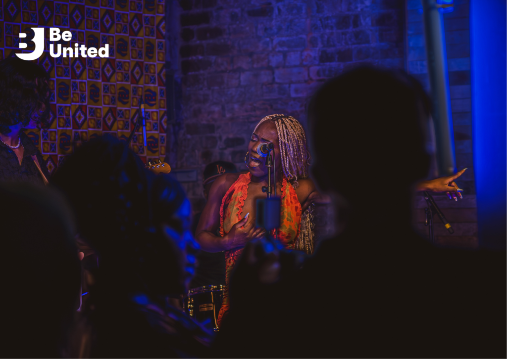 Viv Latifa, a Black woman with long light braids, wearing an orange dress, singing on stage. The audience is visible in the foreground.