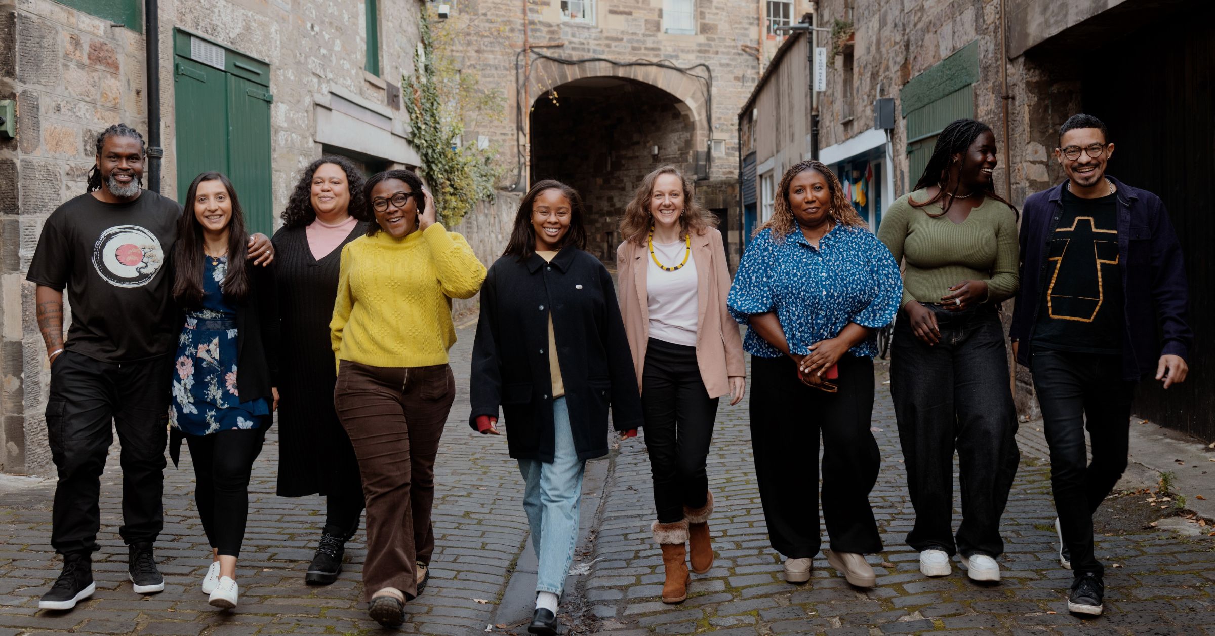 Nine members from the Be United team and Board walk together down a cobbled street, smiling. They are dressed casually and warmly, with Edinburgh's stone buildings in the background.