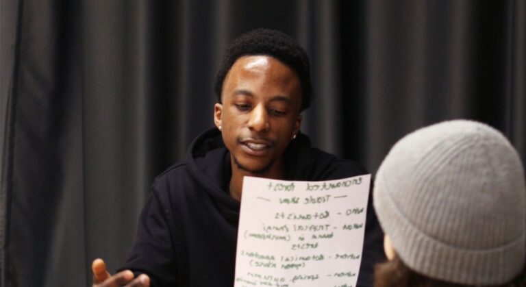 A young Black man sits indoors holding a handwritten planning sheet while speaking to another person, capturing a moment of collaborative discussion during a training session.